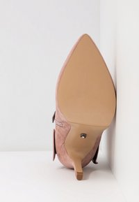 Chaussure à talon haut en daim rose à bout pointu vue par la semelle, montrant un talon étroit et une semelle extérieure beige lisse sur fond blanc.