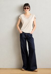 Beige short-sleeve top met een v-hals en kantdetails, gecombineerd met donkerblauwe flared denim jeans en tan schoenen. Zachte texturen en casual pasvorm.