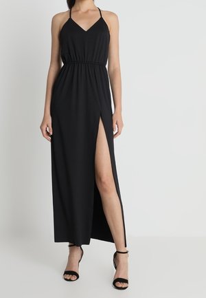 Schwarzes Maxikleid mit tiefem V-Ausschnitt und dünnen Trägern, mit einem elastischen Taillenbund und einem hohen Schlitz am Oberschenkel. Kombiniert mit schwarzen Sandalen mit Absatz.