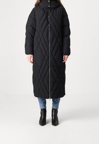 Lange zwarte pufferjas met een diamanten quiltpatroon, hoge kraag en een rits aan de voorkant. Gedragen met blauwe jeans en enkellaarsjes.