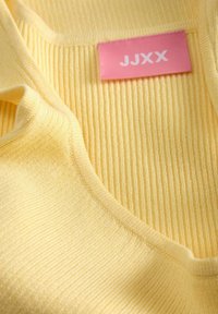 Κίτρινο πλεκτό ύφασμα με ραβδώσεις και ένα ροζ ορθογώνιο ετικέτα με το κείμενο "JJXX" κοντά στη λαιμόκοψη.