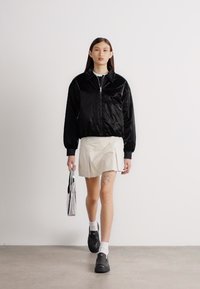 Jachetă bomber neagră și lucioasă cu fermoar, asortată cu o fustă bej deschis, sosete până la gleznă și pantofi negri; ținând o geantă metalică.