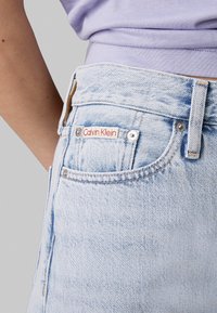 Vaqueros azul claro con un acabado desgastado, que cuentan con una etiqueta roja y blanca de Calvin Klein en el bolsillo delantero y herrajes metálicos plateados.