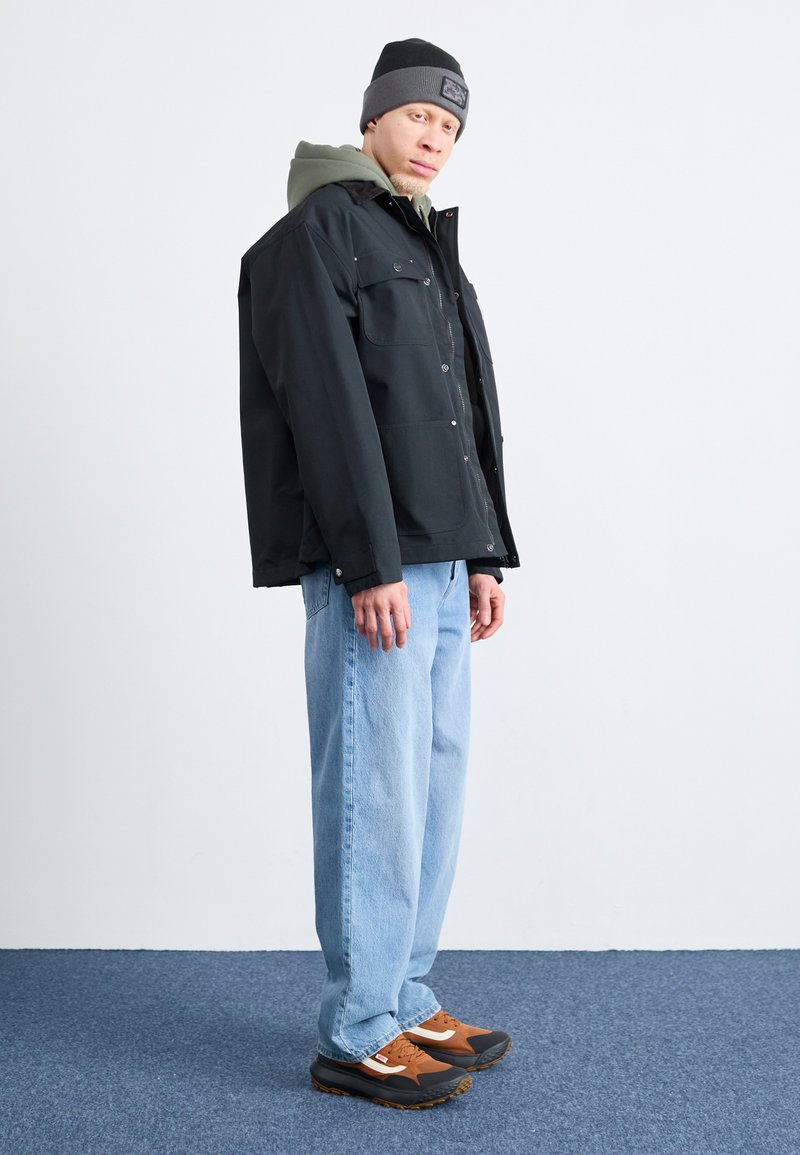 Giubbotto nero con due tasche sul petto, indossato sopra un felpa verde. Jeans azzurri chiari e scarpe da ginnastica marroni con dettagli grigi. Berretto grigio in testa.