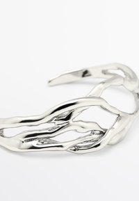 Bracciale in metallo argentato con un design aperto astratto e organico che ricorda rami intrecciati su uno sfondo bianco.