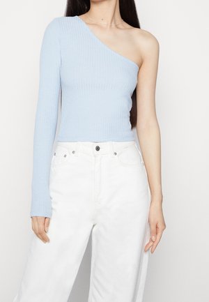 Mujer vistiendo un top azul claro de un solo hombro con textura acanalada, manga larga en el brazo izquierdo y pantalones blancos de tiro alto.