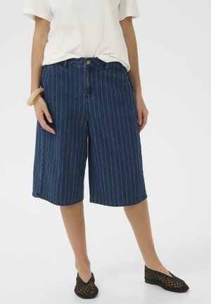Frau trägt marineblaue, knielange Shorts mit breiten Beinen und Nadelstreifen, weißes kurzärmeliges Oberteil, schwarze geflochtene Ballerinas und beiges Armband, steht still.