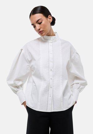 Blouse en coton blanc avec un col montant, devant boutonné et plis structurés le long du centre ; manches bouffantes avec poignets froncés.