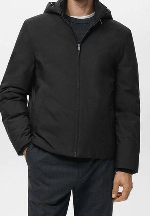 Mies, jolla musta vetoketjullinen hupparintakki, kädet taskuissa, tummien housujen ja kerrostetun neulepaidan päällä.