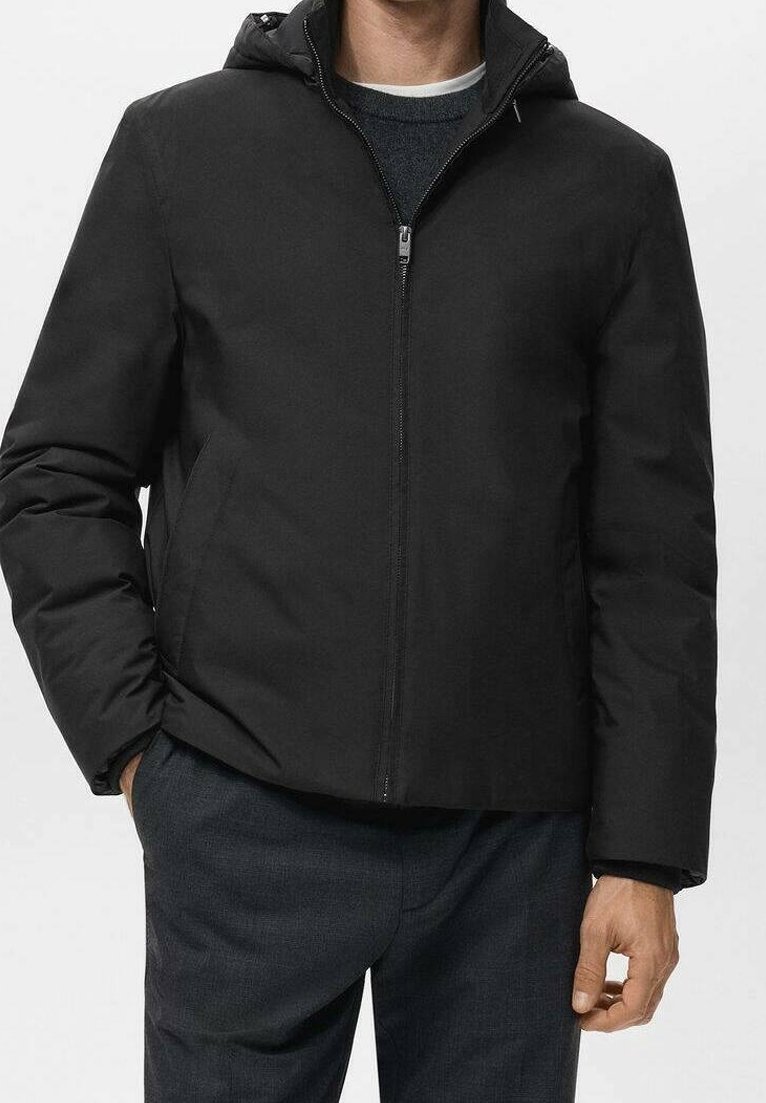 Homme portant une veste à capuche noire zippée, les mains dans les poches, sur un pantalon sombre et un pull en maille superposé en dessous.