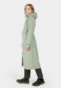 Impermeable largo de color verde claro con capucha, puños ajustables y cremallera frontal. Llevado sobre pantalones negros y zapatillas deportivas negras.