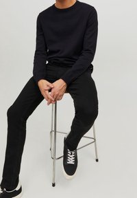 Maglione nero a maglia con scollatura rotonda, abbinato a jeans neri aderenti e sneakers nere con lacci bianchi, seduto su uno sgabello di metallo.