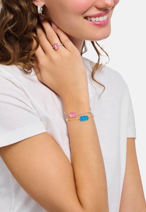 Lächende Person mit silbernen Creolen, einem pinkfarbenen Gummibärchen-Ring und passenden pinken und blauen Gummibärchen-Armbändern auf einem weißen Hemd.