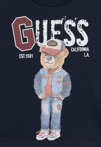 Donkerblauwe t-shirt met een graphic van een beer in een spijkerjack en pet, met de tekst "GUESS CALIFORNIA L.A. EST.1981" prominent weergegeven.