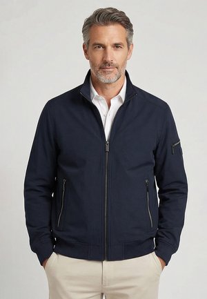 Man van middelbare leeftijd met zout-en-peper haar draagt een marineblauw jasje met rits over een wit overhemd met kraag, staand met handen in de zakken.