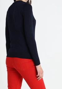 Frau in einem eng anliegenden, langen navy Pullover und hellroten Jeans, die mit leicht zur Kamera gewandtem Rücken steht.