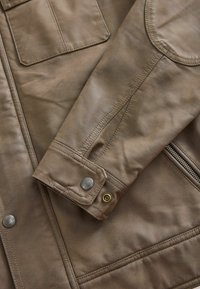 Chaqueta de cuero marrón con cierre de cremallera, bolsillo en el pecho y botones a presión en los puños. Textura suave con detalles de costura visibles.