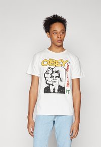 Λευκό βαμβακερό T-shirt με γραφιστικό σχέδιο που απεικονίζει μια γροθιά και ένα πρόσωπο, με το κείμενο "ΥΠΑΚΟΥΣΤΕ ΝΟΜΙΜΟΠΟΙΗΣΤΕ ΤΟ" σε κίτρινο, κόκκινο και πράσινο χρώμα.