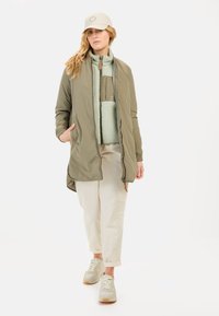 Khaki gewatteerde lange jas met ribbels aan de mouwen, gelaagd met een lichtgroene fleece voering. Gecombineerd met crèmekleurige broek en grijze sneakers.