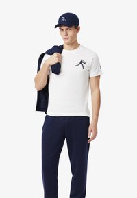 Camiseta blanca con logo azul oscuro, pantalones deportivos azul marino, gorra azul marino y chaqueta oscura colgada sobre el hombro, con una textura suave y un corte relajado.
