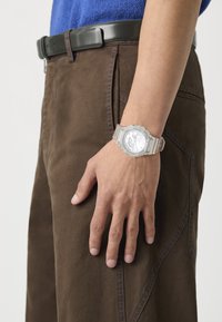 Transparent resin watch with a round silver face and digital display, worn on a hand resting against brown pants with blue stitching.