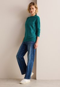 Teal stickad tröja med hög hals och sömdetaljer, ihopparad med löst sittande blå jeans och vita sneakers på en neutral bakgrund.