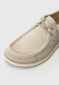 Beige og grå suedeskole med snøredesign, som har en avrundet tå og kontrasterende søm langs kantene. Glatt tekstur.