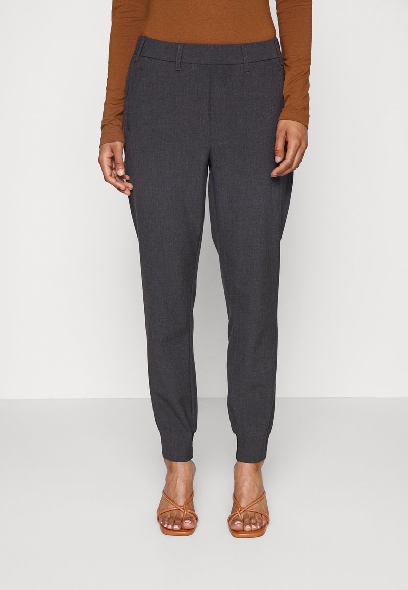 Personne portant un pantalon gris foncé fuselé, un haut marron à manches longues et des sandales marron à brides ouvertes à l'avant, debout sur un fond blanc.