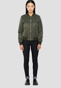 Olijfgroene bomberjack met een ritssluiting aan de voorkant, geribbelde manchetten en zoom, en zijzakken, gecombineerd met donkere jeans en zwarte schoenen.