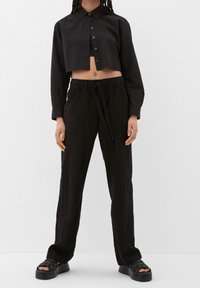Chemise noire courte à boutons avec col, associée à un pantalon noir ample avec taille élastique et poches. Sandales plateformes noires.