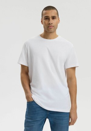 Mężczyzna o krótkich włosach, ubrany w gładką białą koszulkę z krótkim rękawem i niebieskie dżinsy, stoi z jedną ręką w kieszeni na jednolitym tle.