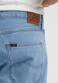 Blå denimjeans har en brun läderetikett på midjebandet med "Lee" präglat, samt en liten etikett på bakfickan med orange sömnad.