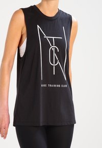 Top sin mangas negro de Nike Training Club con letras abstractas blancas, usado sobre un sujetador deportivo blanco, combinado con leggings negros.