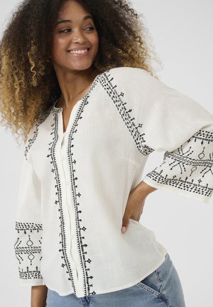 Blouse en lin blanc avec des motifs brodés noirs le long des manches et à l'avant. Dotée d'un décolleté en V et de boutons au centre.