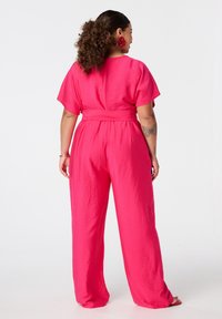 Combinaison rose vif avec des manches courtes, jambes larges et taille cintrée. Tissu lisse et texturé avec un subtil éclat. Détails élégants à l'arrière.