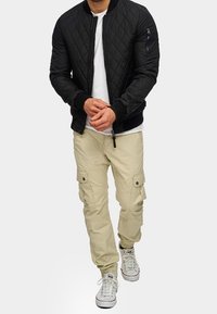 Czarna pikowana kurtka typu bomber z prążkowanym kołnierzem i mankietami, w zestawie z beżowymi spodniami cargo z bocznymi kieszeniami oraz białymi trampkami.