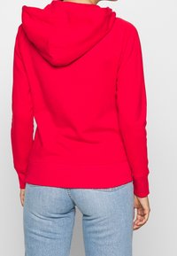 Sudadera roja con capucha, de mangas largas, puños y bajo de ribete. Textura de tela suave. Combinada con jeans de color azul claro.