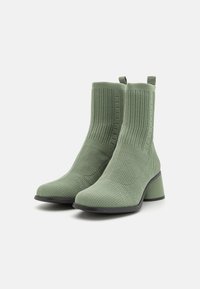 Botas de tobillo de punto verde con tacón cuadrado, que presentan una superficie texturizada y lengüetas para facilitar el calzado. La suela es de color negro, contrastando con la parte superior.