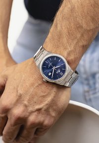 Reloj de acero inoxidable plateado con esfera azul y función de fecha, con un bisel texturizado y una correa integrada en una muñeca.