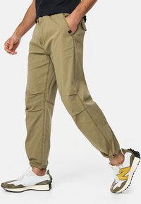 Khaki cargo pants with a relaxed fit, elastic cuffs, side pockets, and contrast stitching. Paired with white and grey sneakers.