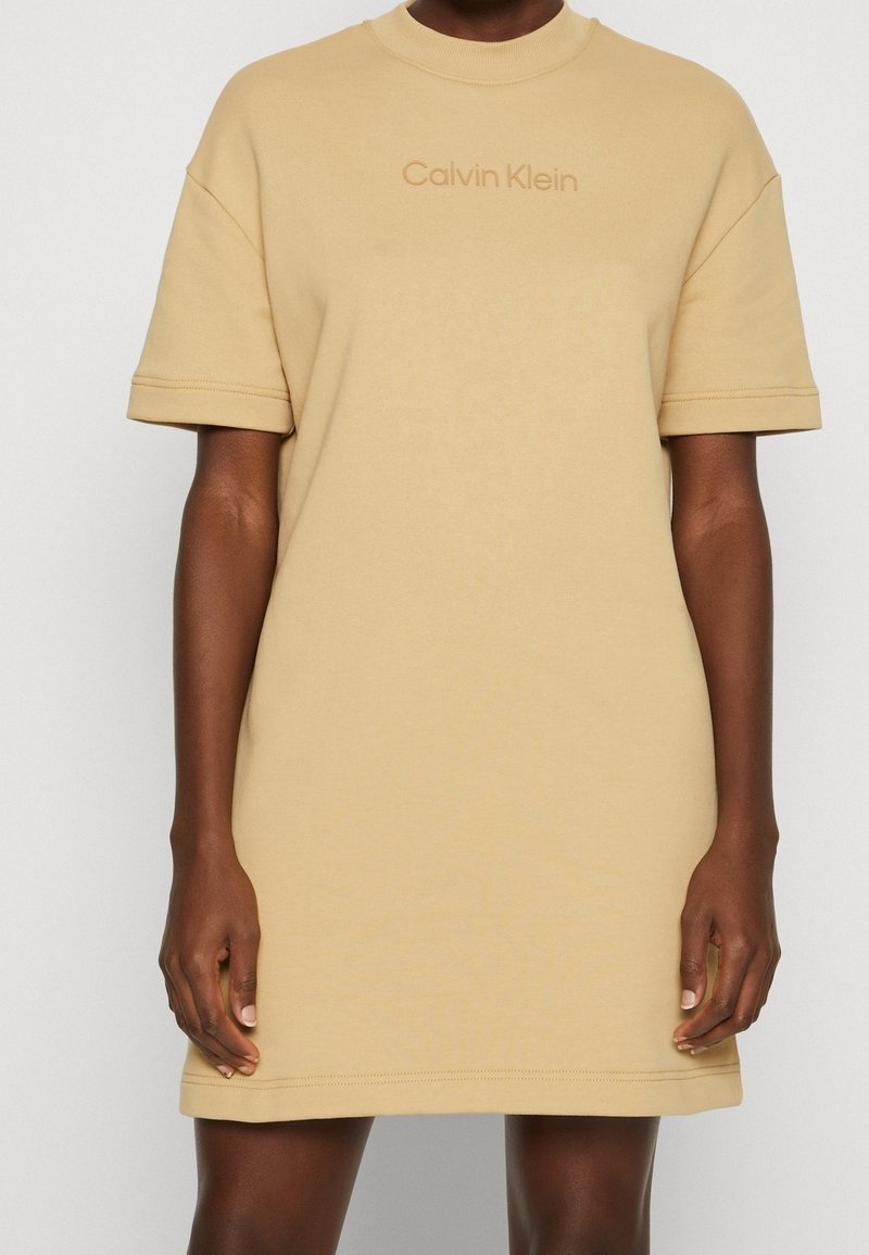Robe beige Calvin Klein à manches courtes portée par une personne, avec un col rond haut et un logo brodé sur la poitrine.