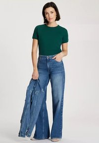 Zielona koszulka z krótkim rękawem w zestawie z niebieskimi dżinsami o podwyższonym stanie i szerokich nogawkach. Niebieska dżinsowa kurtka jest zarzucona na jej ramię.