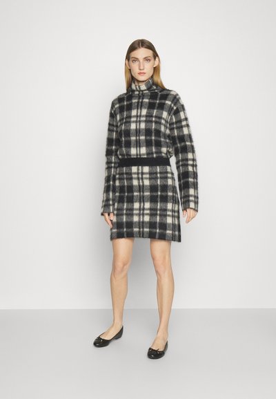 Ženska oblečena v dolgo črno-belo karirano obleko s turtleneck ovratnikom in črnimi baletnimi čevlji, ki stoji ob beli steni.