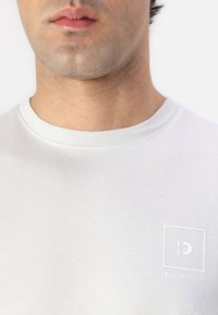 Hvit bomulls t-skjorte med rund hals og korte ermer, med en subtil brodert logo i en firkant på venstre brystområde.