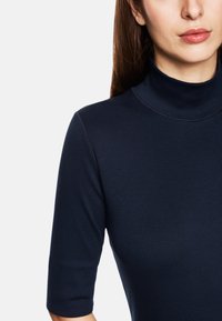 Vrouw met een marineblauwe coltrui met mouwen tot de elleboog, weergegeven van schouders tot kin op een witte achtergrond.