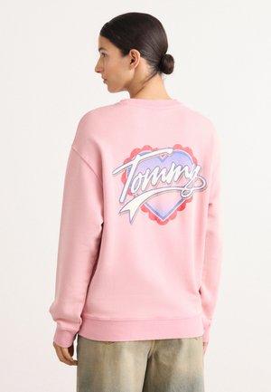 Sudadera rosa con cuello redondo y puños de canalé. Presenta un gran gráfico multicolor en la parte posterior con el nombre "Tommy" dentro de un corazón decorativo.