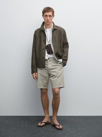 Olivgrüne Jacke über einem weißen Hemd, beigen Shorts und braunen Flip-Flops. Merkmale eine kleine braune Umhängetasche um den Hals.