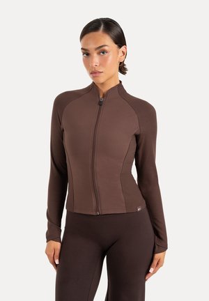 Femme portant une veste moulante marron zippée et des leggings marron assortis sur un fond clair uni.