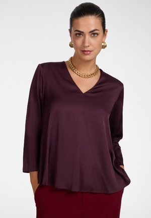 Vrouw met donker haar in een gladde knot, gekleed in een paarse blouse met V-hals, rode broek, gouden ketting en gouden oorringen tegen een witte achtergrond.