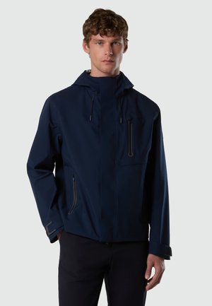 Mann trägt eine marineblaue Kapuzenjacke mit Reißverschlusstaschen an Brust und Seiten, steht vor einem schlichten hellen Hintergrund.